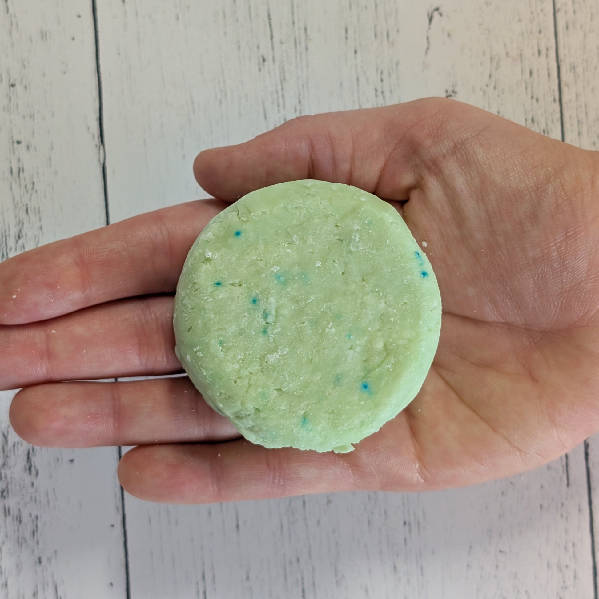Hand holding a green bath bomb with blue specks on a light wooden surface