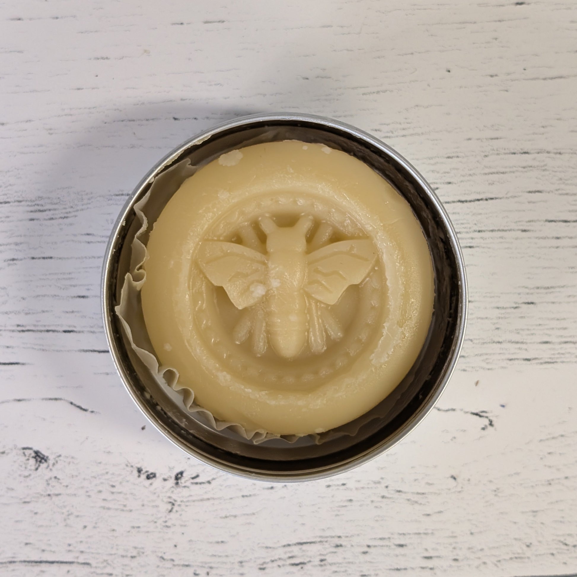 Beige soap with bee design in a metal tin on a white wooden surface